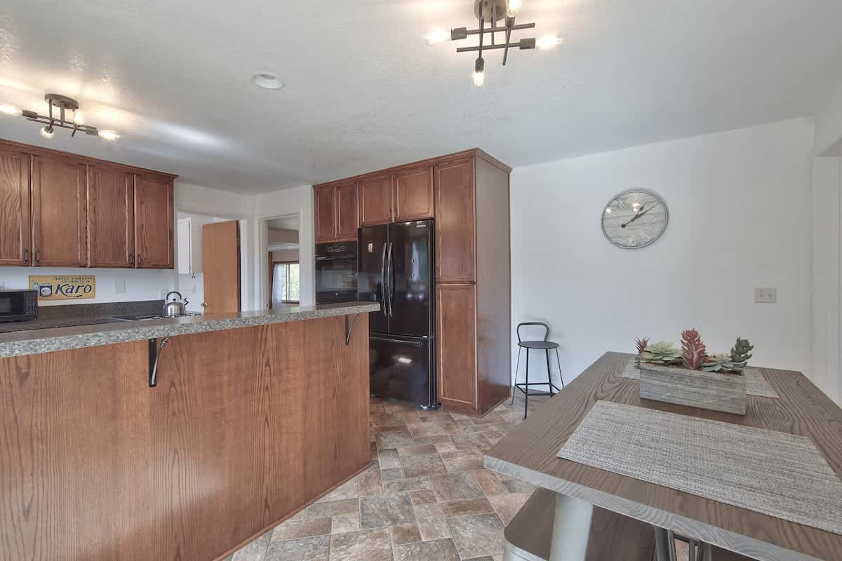An expansive view of the kitchen and dining area, featuring dark wood cabinets, dark countertops, and a central island with a breakfast bar. A dining table with stools is visible, and the space is illuminated by modern light fixtures and recessed lighting.