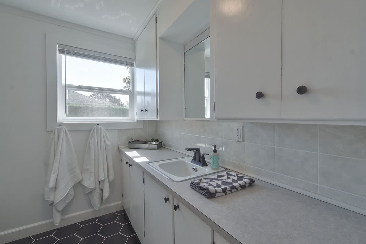 Functional laundry room with white upper and lower cabinets, a utility sink set in a light gray countertop, and dark hexagonal floor tiles. A window provides natural light.
