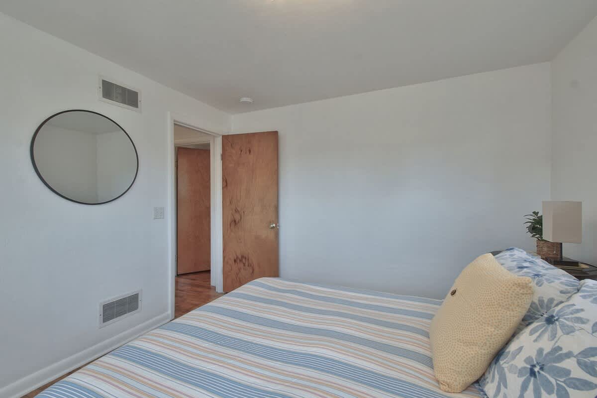 Another view of bedroom-2, highlighting the warm wooden door and a round black-framed mirror on the white wall. The bed with striped bedding is partially visible, and hardwood floors extend throughout the room.