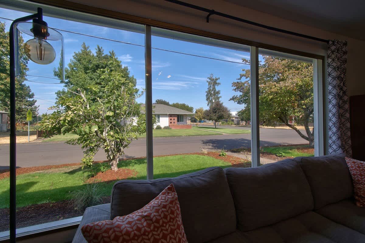 Panoramic view from the living room's large window, showcasing a residential street with green lawns, mature trees, and houses under a bright blue sky.