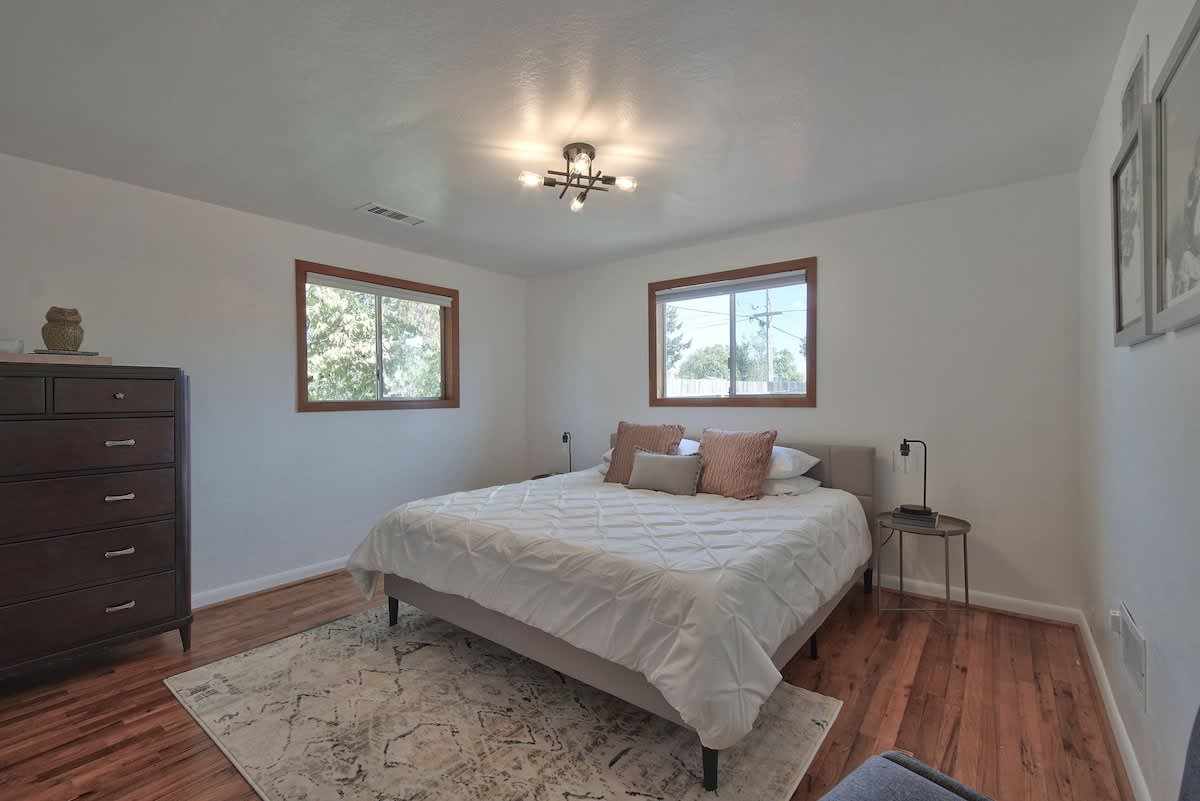 Spacious bedroom with a queen-sized bed, light gray upholstered frame, white bedding, and a patterned rug on hardwood floors. Two windows with wooden frames overlook the exterior.