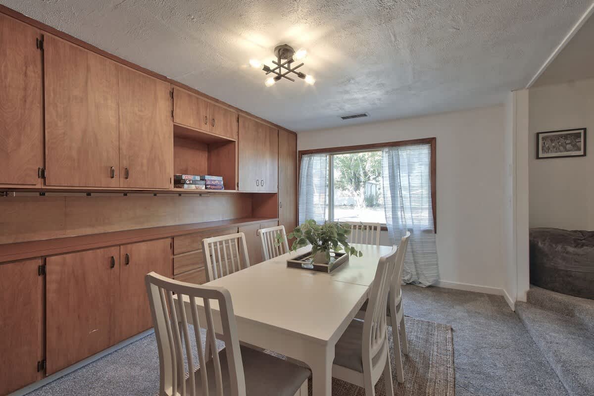 Inviting dining space with a white table and chairs, flanked by a wall of warm wooden built-in cabinets and shelving, and illuminated by natural light from a window with sheer patterned curtains.