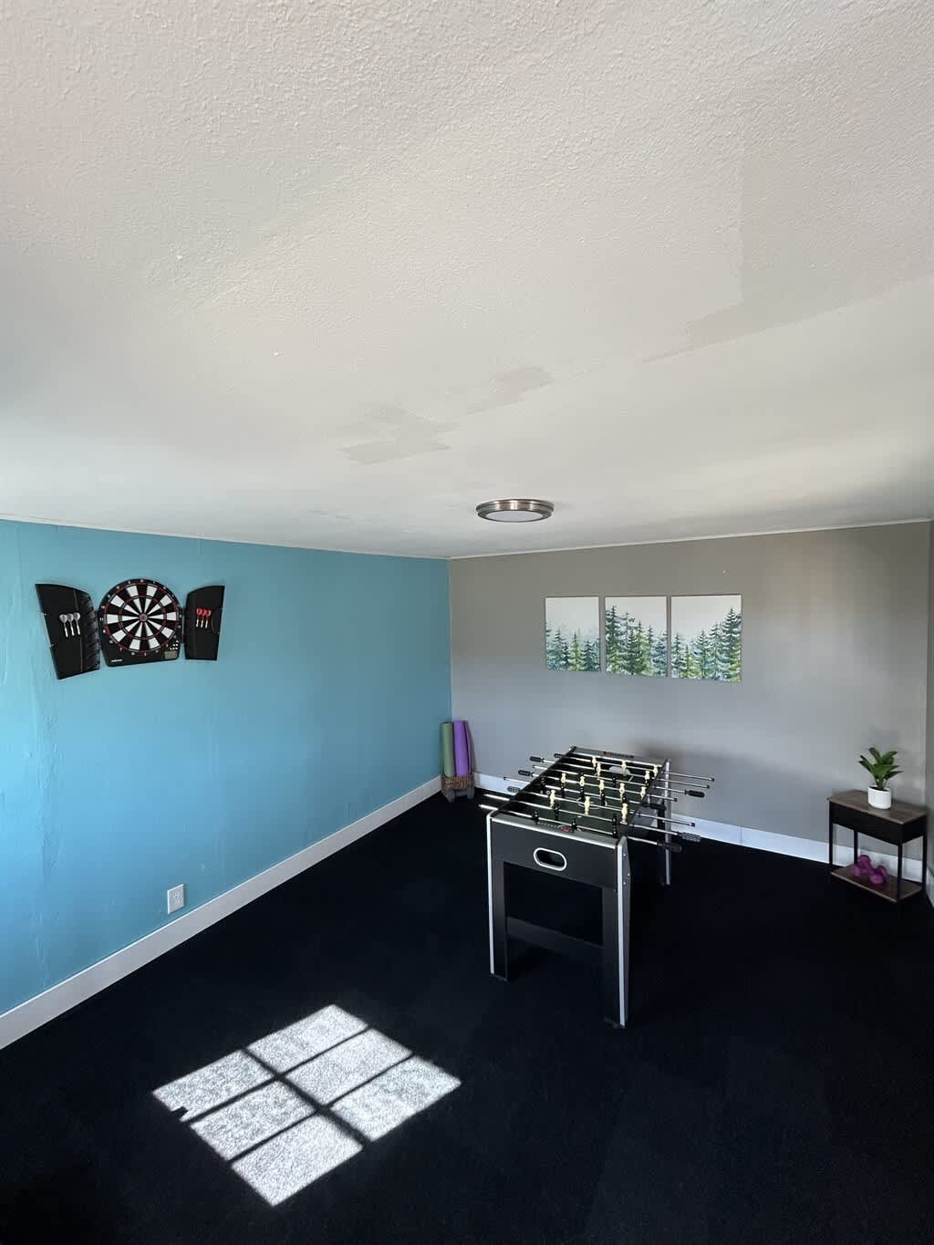 A spacious game room features a dartboard on a vibrant blue accent wall and a foosball table on dark carpet, with three framed nature prints on the adjacent gray wall.
