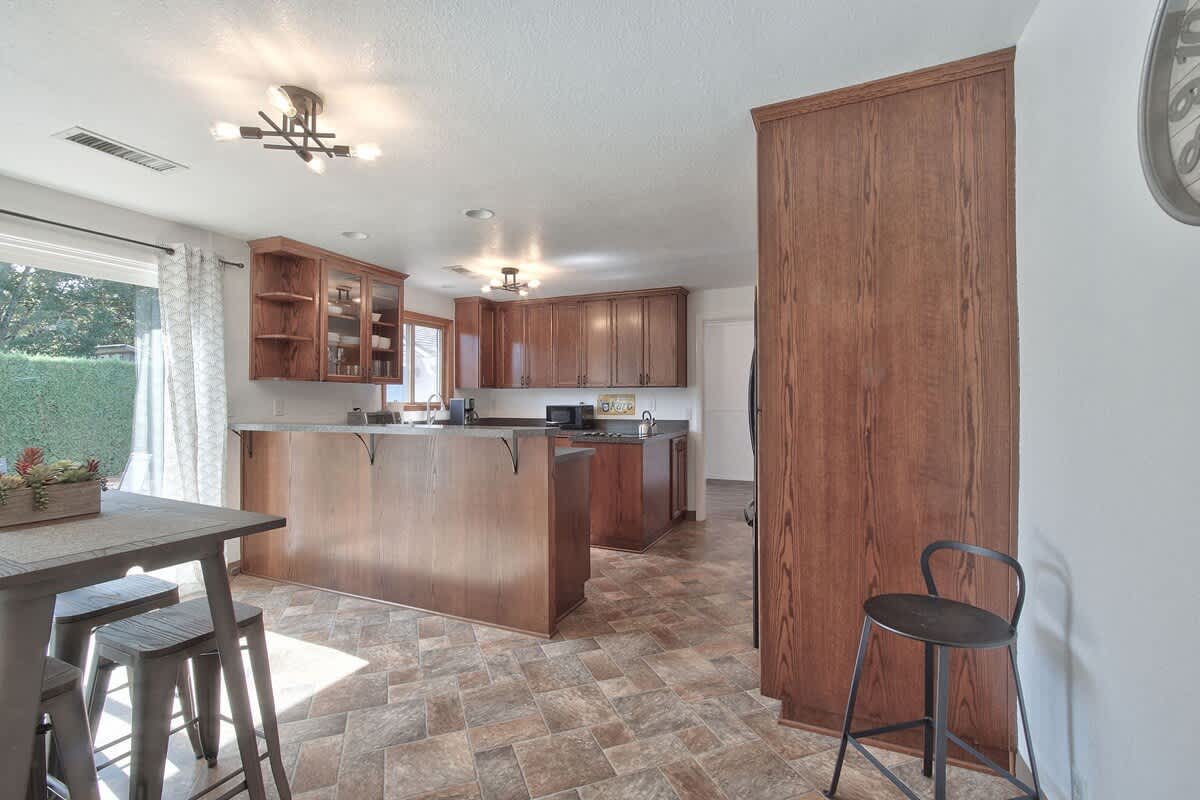 An open-concept kitchen and dining space featuring dark wood cabinetry, dark countertops, and a kitchen island with a breakfast bar. A long dining table with stools is visible, and a sliding glass door leads to the backyard.
