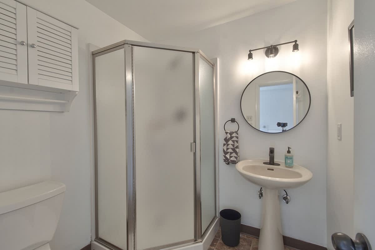 A bright white bathroom featuring a corner shower with frosted glass doors, a white pedestal sink with a round mirror and modern vanity light, and a white toilet. The floor has brown and beige patterned tiles.