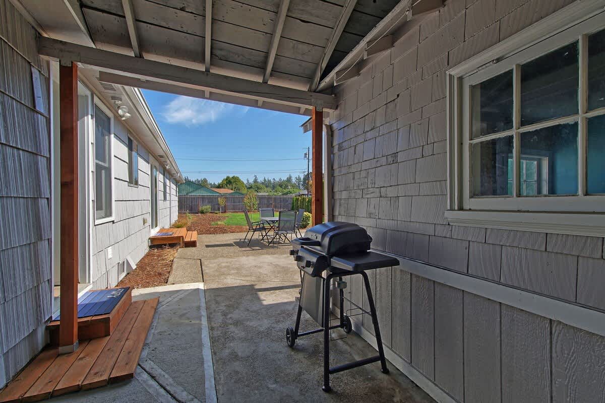 Covered outdoor area with a concrete path leading to the main patio, featuring a black BBQ grill. The side of the house with gray shingle siding and a window is visible, along with wooden steps and posts supporting the roof.