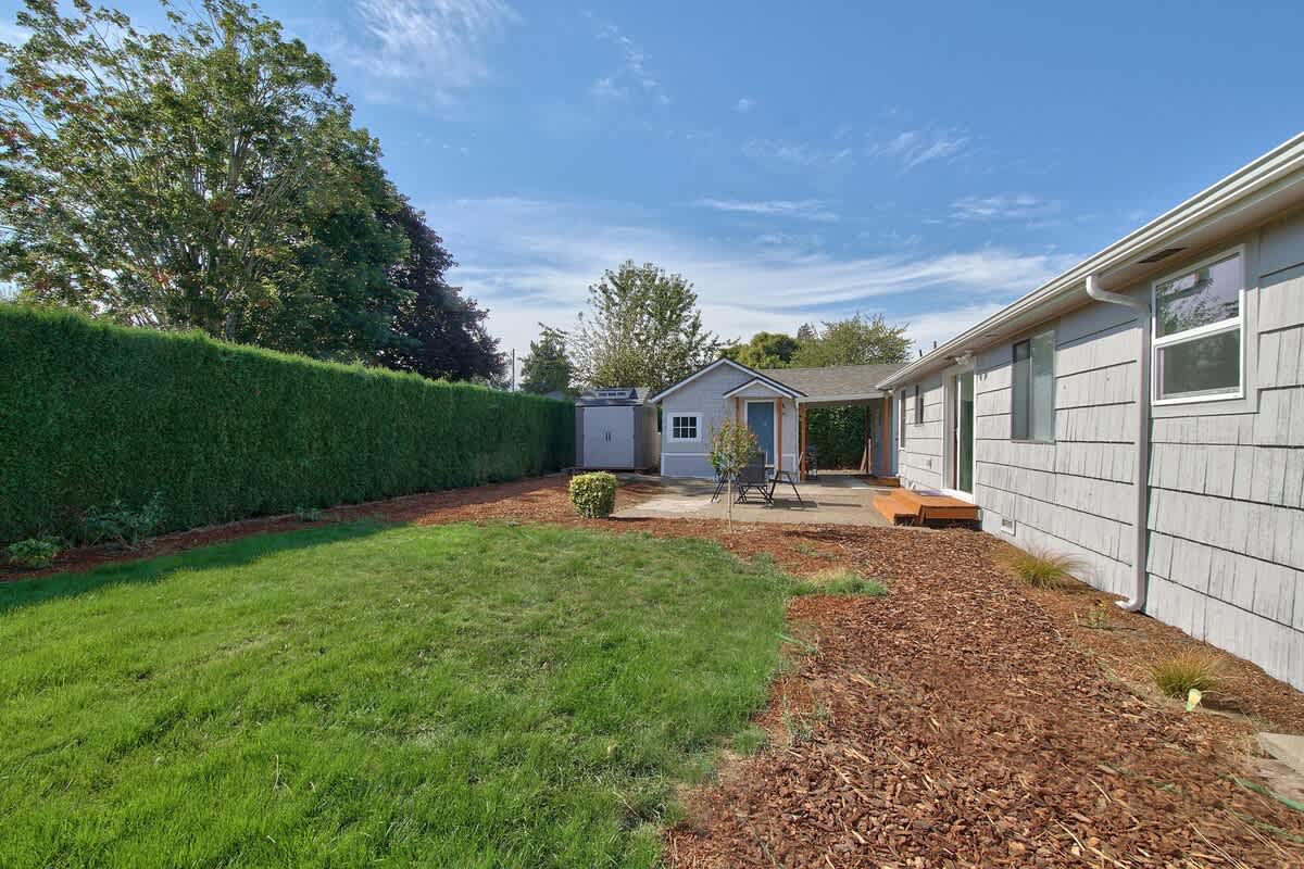 A spacious backyard with a green lawn and mulched areas leads to a small patio with a shed and outdoor seating, bordered by a tall hedge and the house exterior.