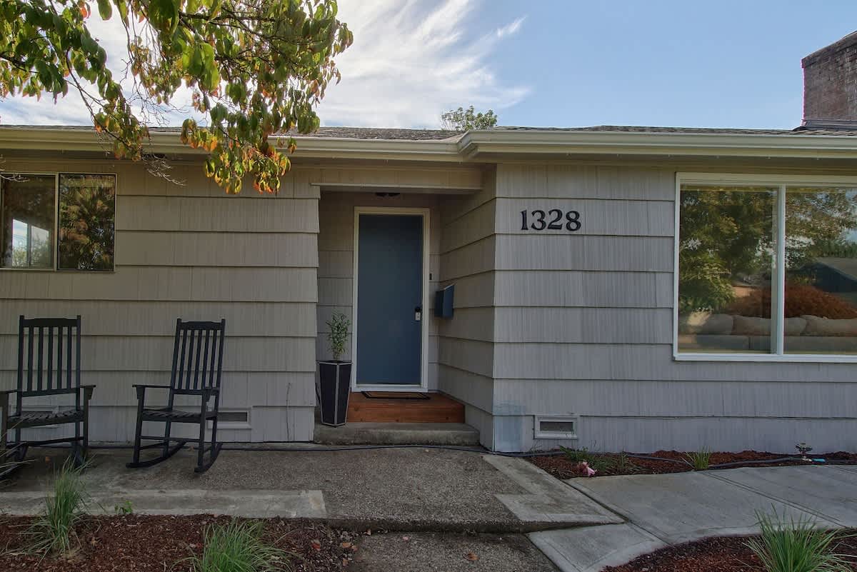 The front entrance of the light-colored shingled house features a dark blue door, house number "1328", and two black rocking chairs on the porch.