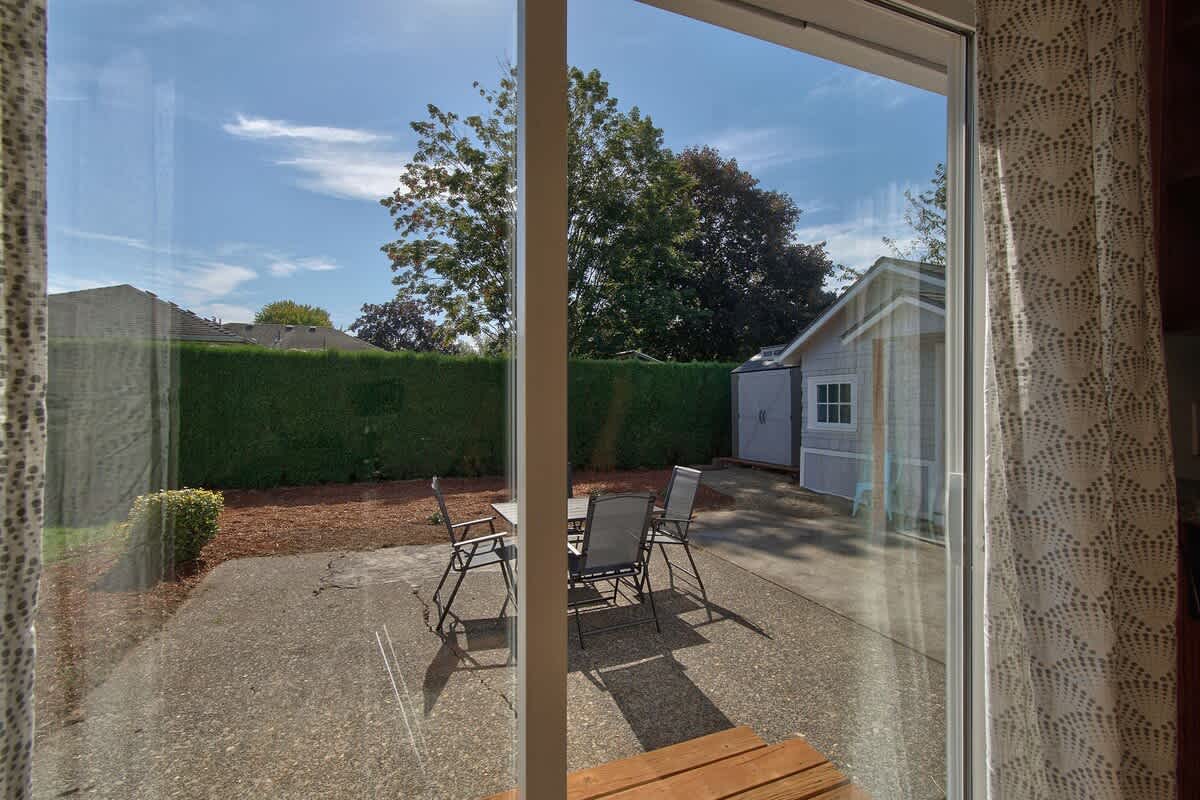 View from inside through a large sliding glass door, looking out onto a concrete patio with outdoor dining furniture. Beyond the patio, a well-maintained green hedge borders the yard under a bright blue sky.