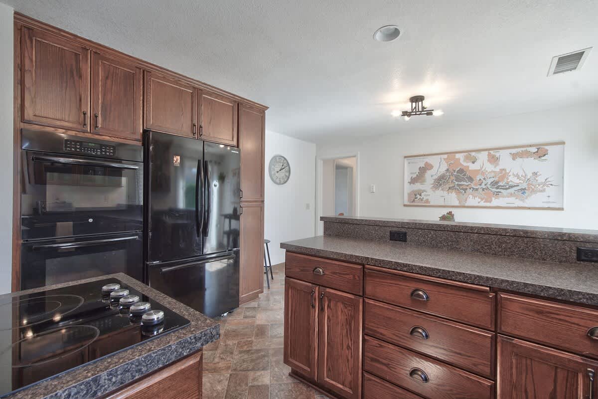 A well-equipped kitchen featuring a built-in black oven and microwave, a black refrigerator, and dark wood cabinetry. The dark speckled countertops provide ample workspace, and a cooktop is integrated into the island.