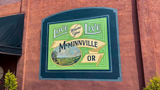 A large, arched green and yellow sign is mounted on a textured reddish-brown wall, featuring the text "LOVE Where You LIVE McMinnville OR" and an oval landscape painting of rolling hills and trees.