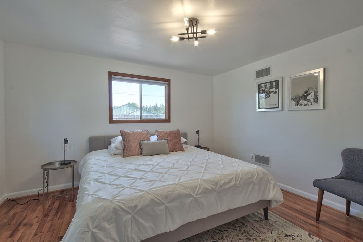 Serene bedroom featuring a large bed with a white quilted duvet and decorative pillows, warm hardwood floors, and a window with a dark wood frame, creating a bright and comfortable retreat.
