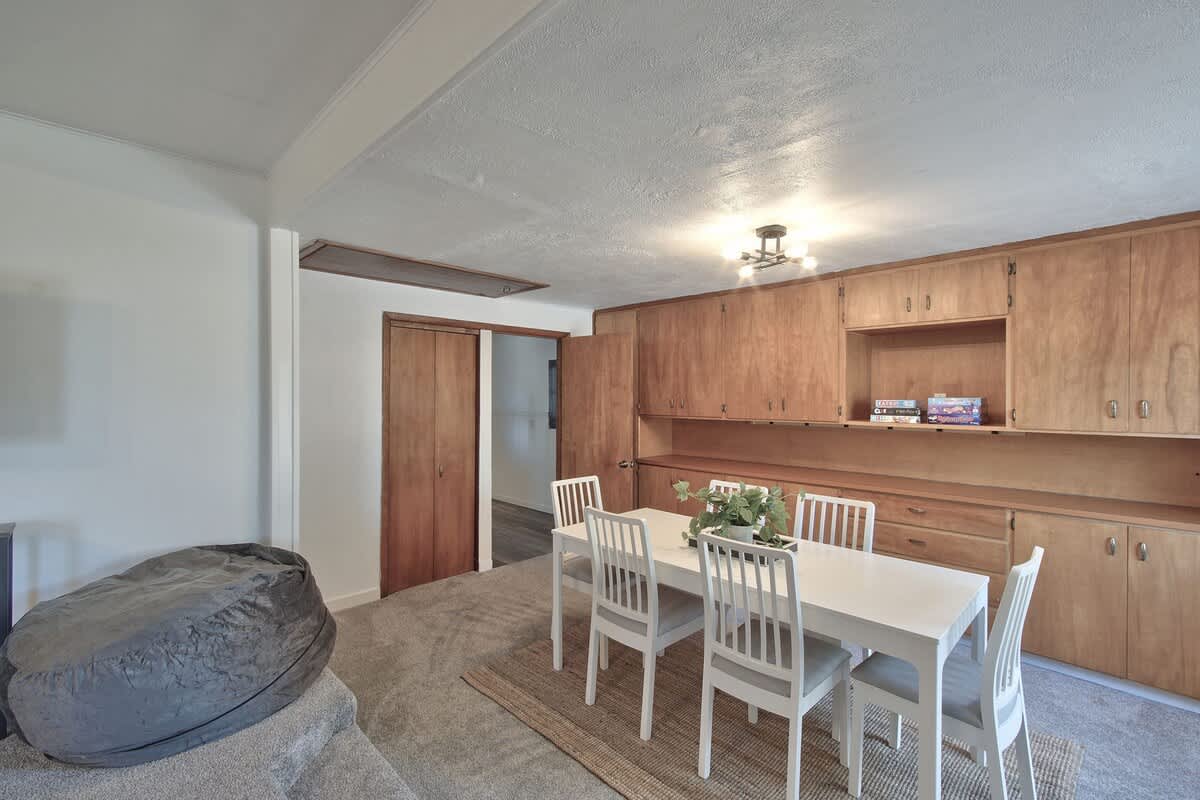 Cozy dining area featuring a white dining table and chairs, complemented by extensive built-in wooden cabinetry and shelving along one wall, with a large grey bean bag chair in the foreground.