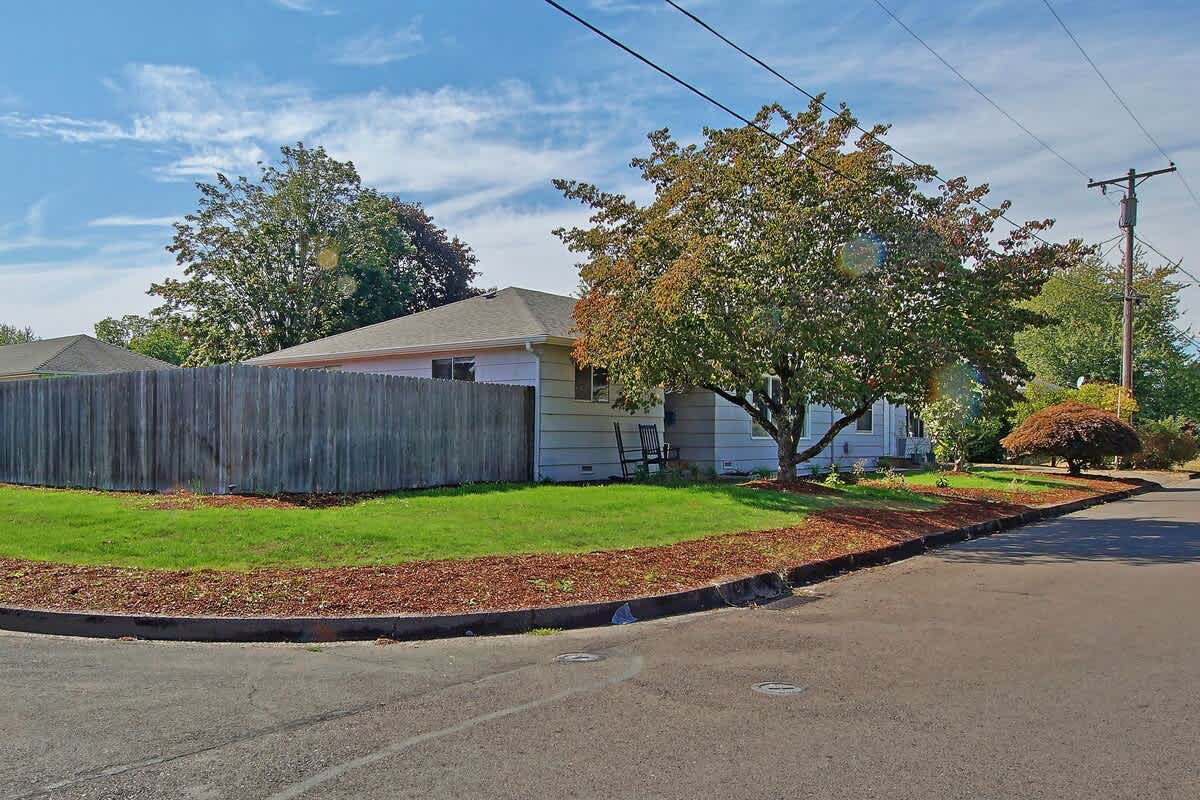 The front exterior of the house, with light-colored siding, is visible from the street corner, featuring a green lawn, mature trees, and a wooden fence.