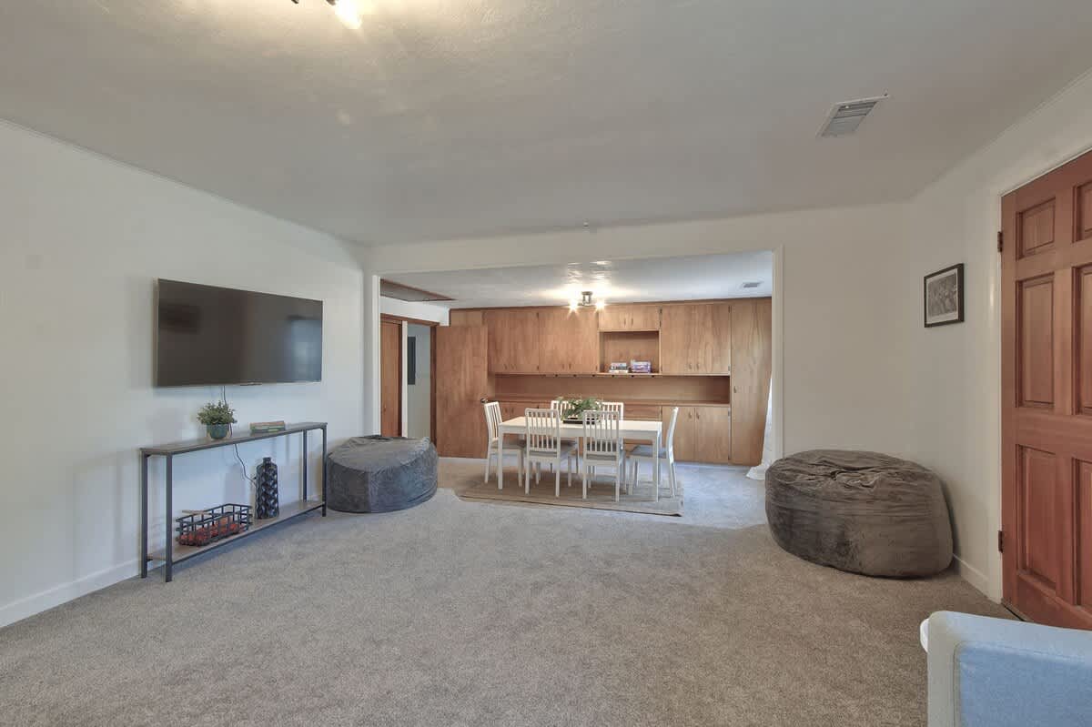 Expansive open-concept living room with light grey carpet, featuring a wall-mounted TV above a console table, two large bean bag chairs, and an open view into the adjacent dining area.
