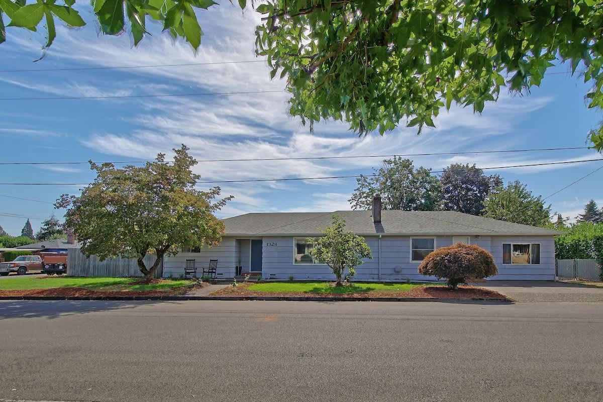 Single-story light gray house with a dark gray roof, visible from the street, surrounded by green lawn and mature trees under a blue sky with white clouds.
