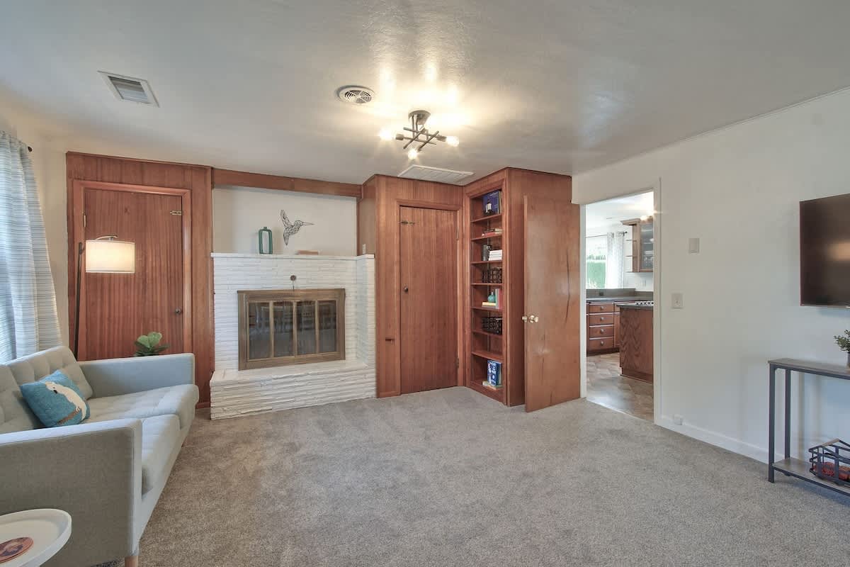 Comfortable living room centered around a white brick fireplace with a wooden mantel, featuring a light grey sofa, warm wooden wall paneling, and a built-in bookshelf.