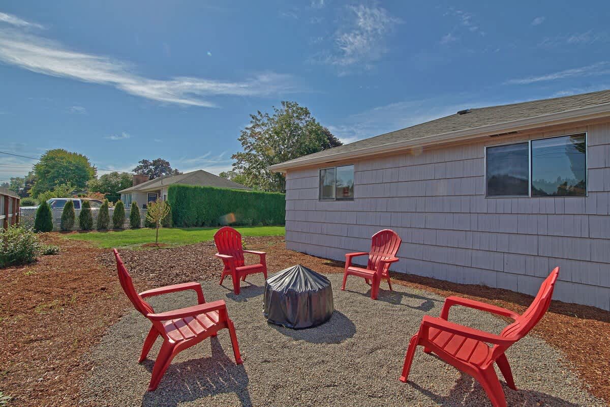 A circular gravel area with a covered fire pit and four red Adirondack chairs sits in a mulched backyard, adjacent to the light-colored shingled house exterior.