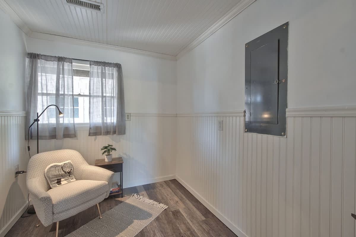 Bright small room with white wainscoting and a comfortable patterned armchair. A small side table with a plant sits next to the chair, and a window with sheer gray curtains allows light into the space.
