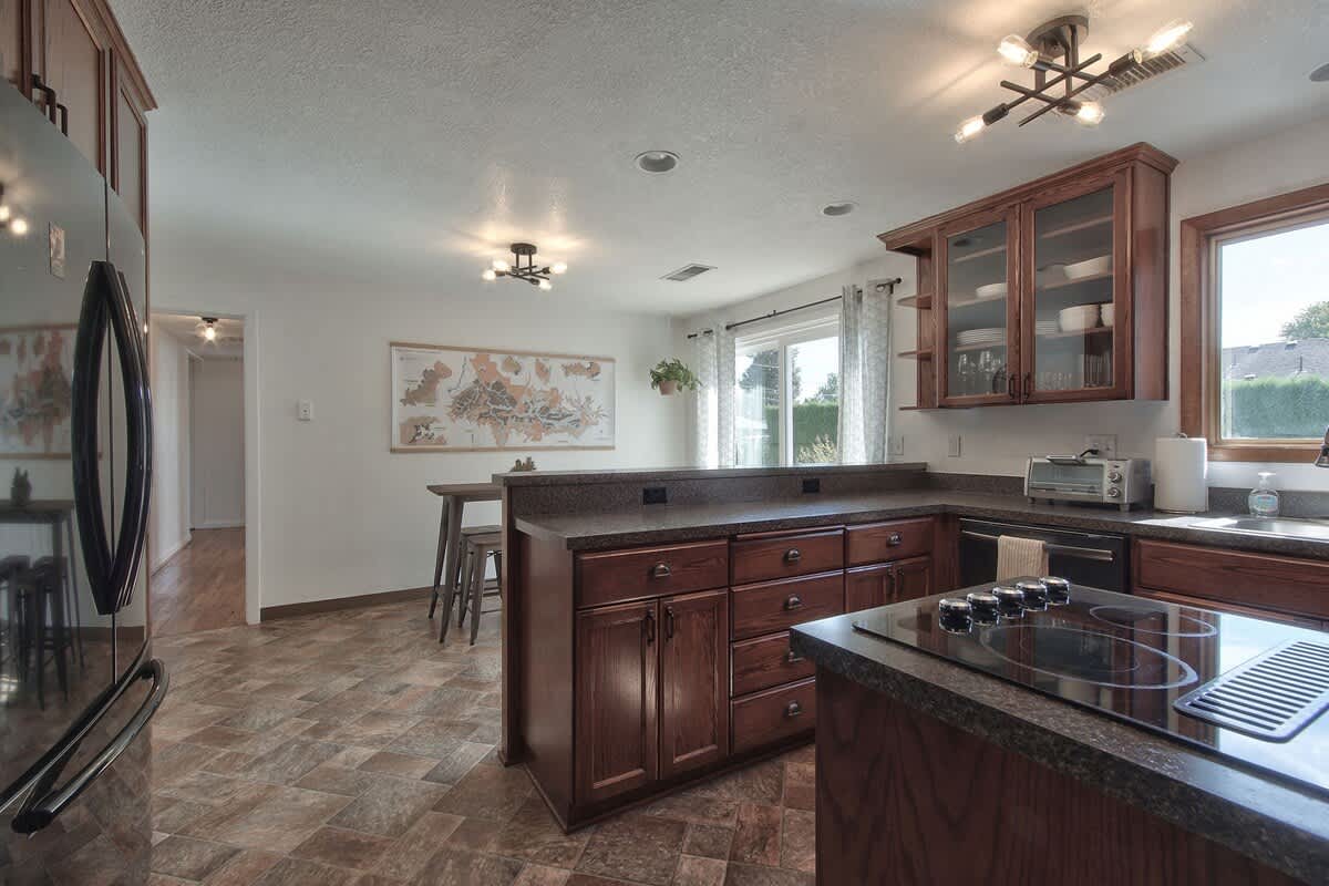 A spacious kitchen featuring a large island with a built-in electric cooktop and dark speckled countertops. Dark wood cabinets and a black refrigerator and oven are visible, with a dining area and sliding glass door in the background.
