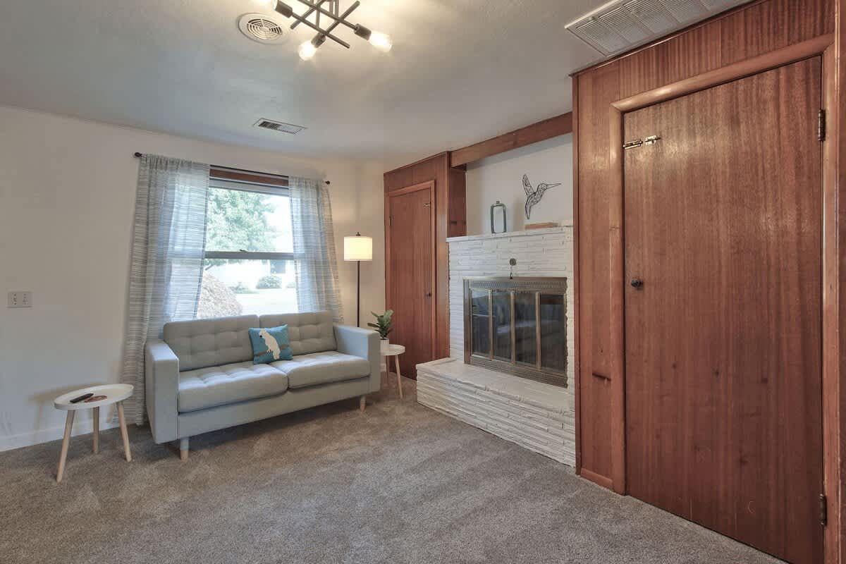 Inviting living room corner showcasing a white brick fireplace with a metal screen, flanked by warm wooden doors, and a light grey sofa positioned by a window with sheer curtains.