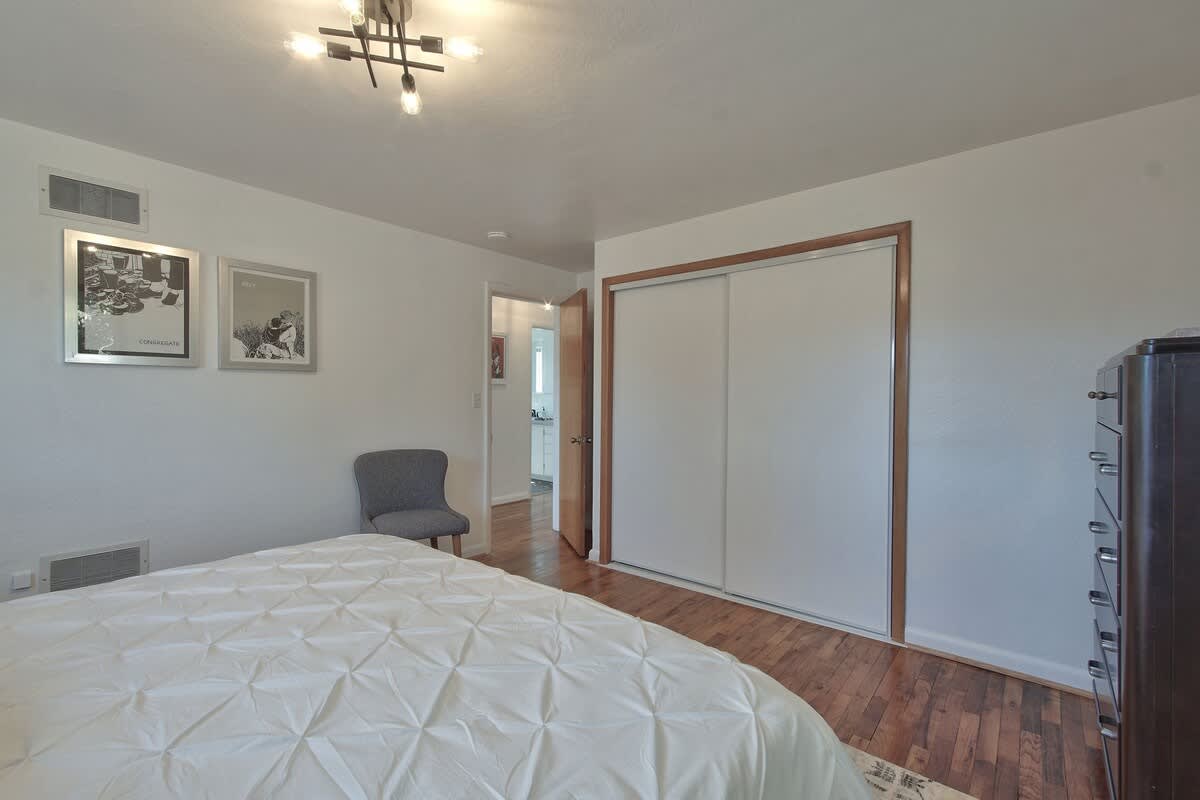 Bright bedroom with white walls, warm hardwood floors, and a bed with white quilted bedding. A modern ceiling light fixture illuminates the space, which also features a closet with white sliding doors and a small gray accent chair.