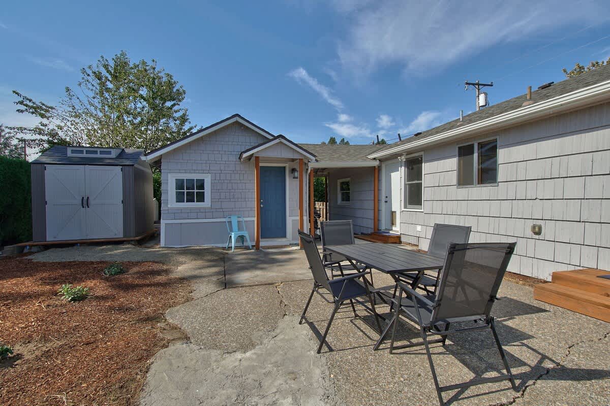 Spacious concrete patio in the backyard featuring a dark gray outdoor dining set with four chairs. A light gray shed with white trim is visible in the background, surrounded by wood chip landscaping and green hedges under a clear sky.