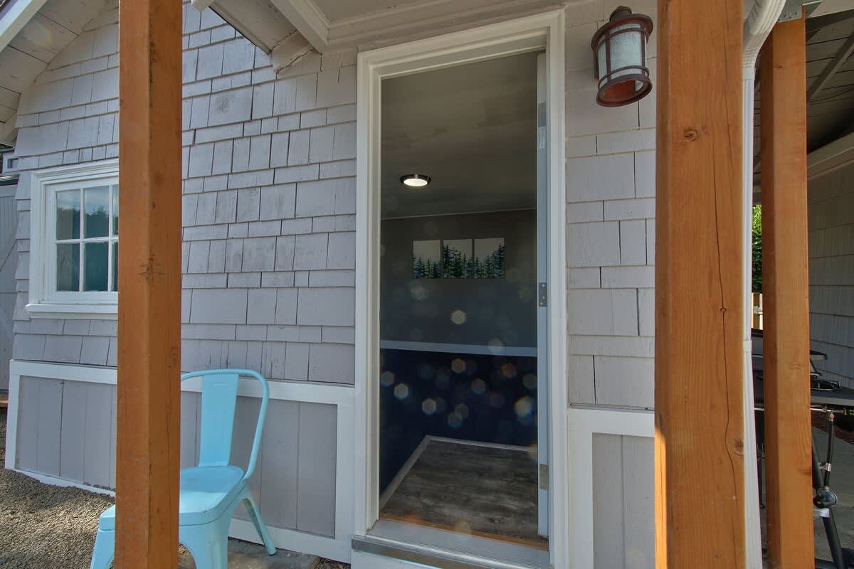 An open glass-paned door reveals a glimpse into an interior room with a dark accent wall, framed by light-colored shingle siding and rustic wooden support beams.