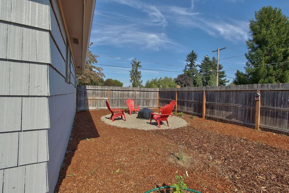A circular gravel fire pit area with four red Adirondack chairs is nestled in a mulched backyard, enclosed by a wooden fence under a bright blue sky.