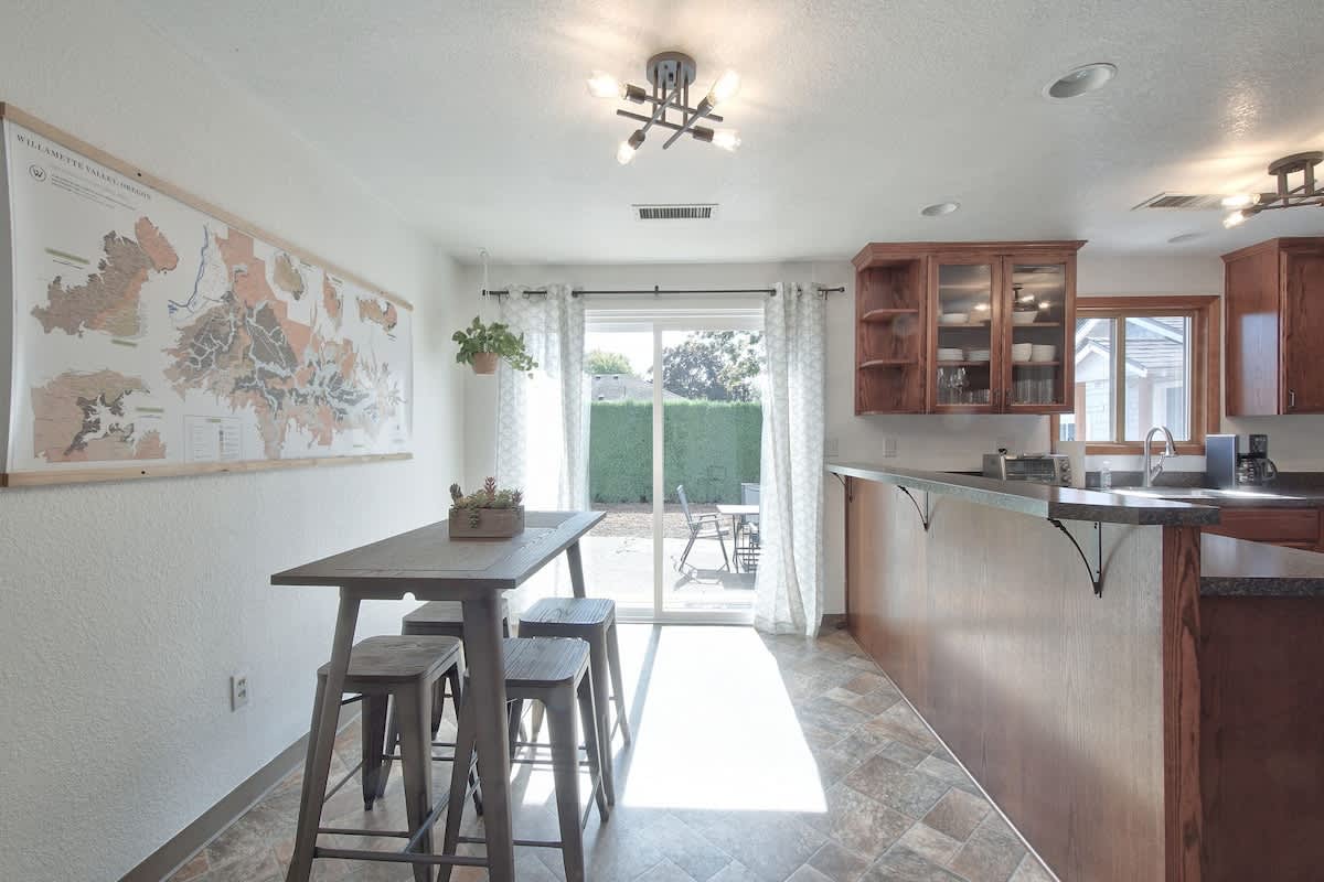 A spacious kitchen and dining area with a long wooden dining table and four metal stools, set against a large map on the wall. A sliding glass door leads to an outdoor patio, and the kitchen features dark wood cabinets and dark countertops.