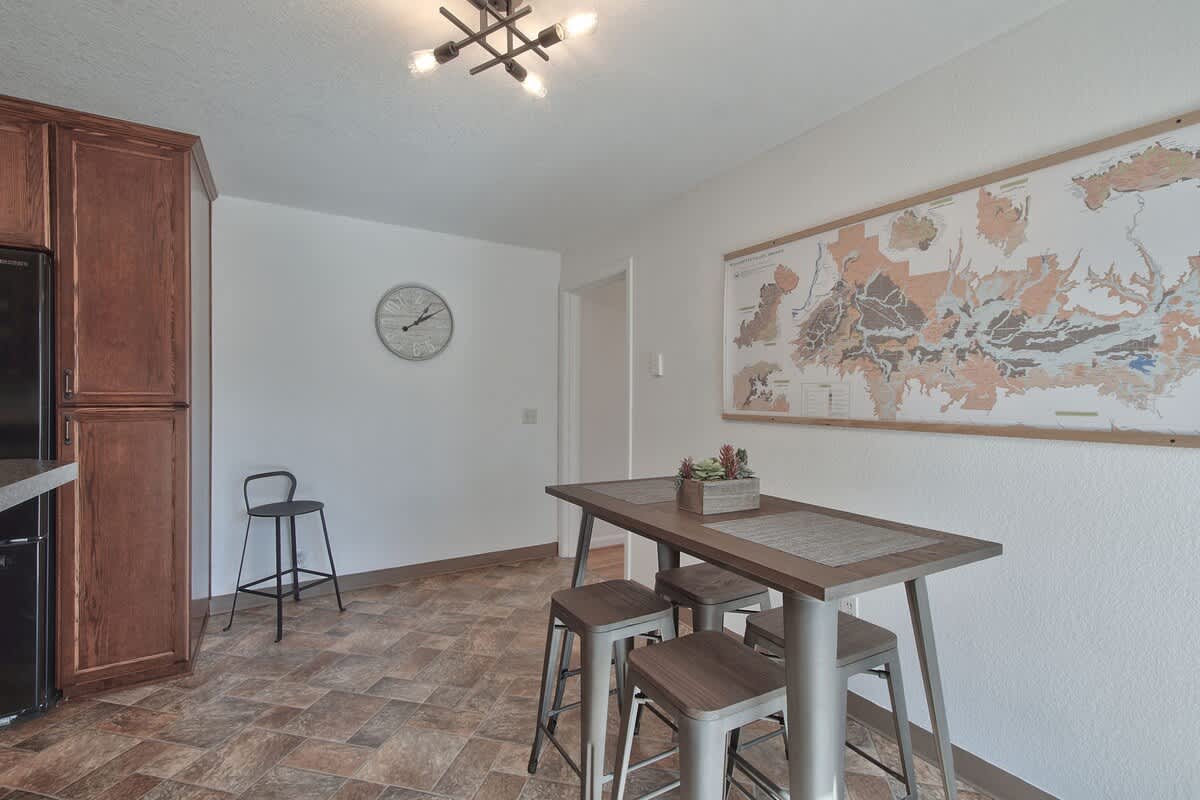 A modern dining area featuring a long rectangular wooden table with four metal stools, positioned beneath a contemporary light fixture. A large map decorates the white wall, and a dark wood cabinet is visible to the left.