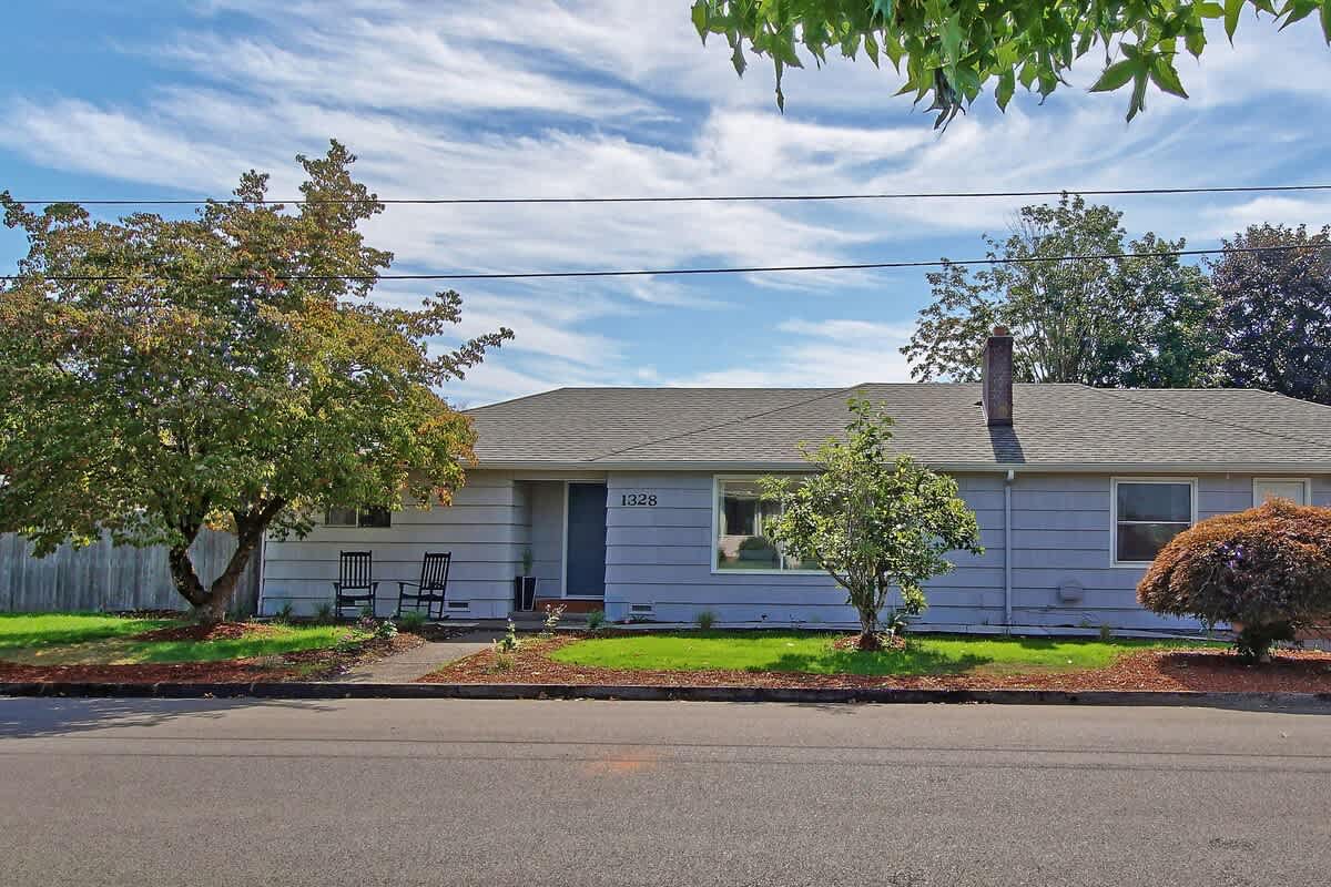 The full front exterior of the single-story house, with light gray shingle siding, is seen from the street, featuring a green lawn, mature trees, and a visible chimney.