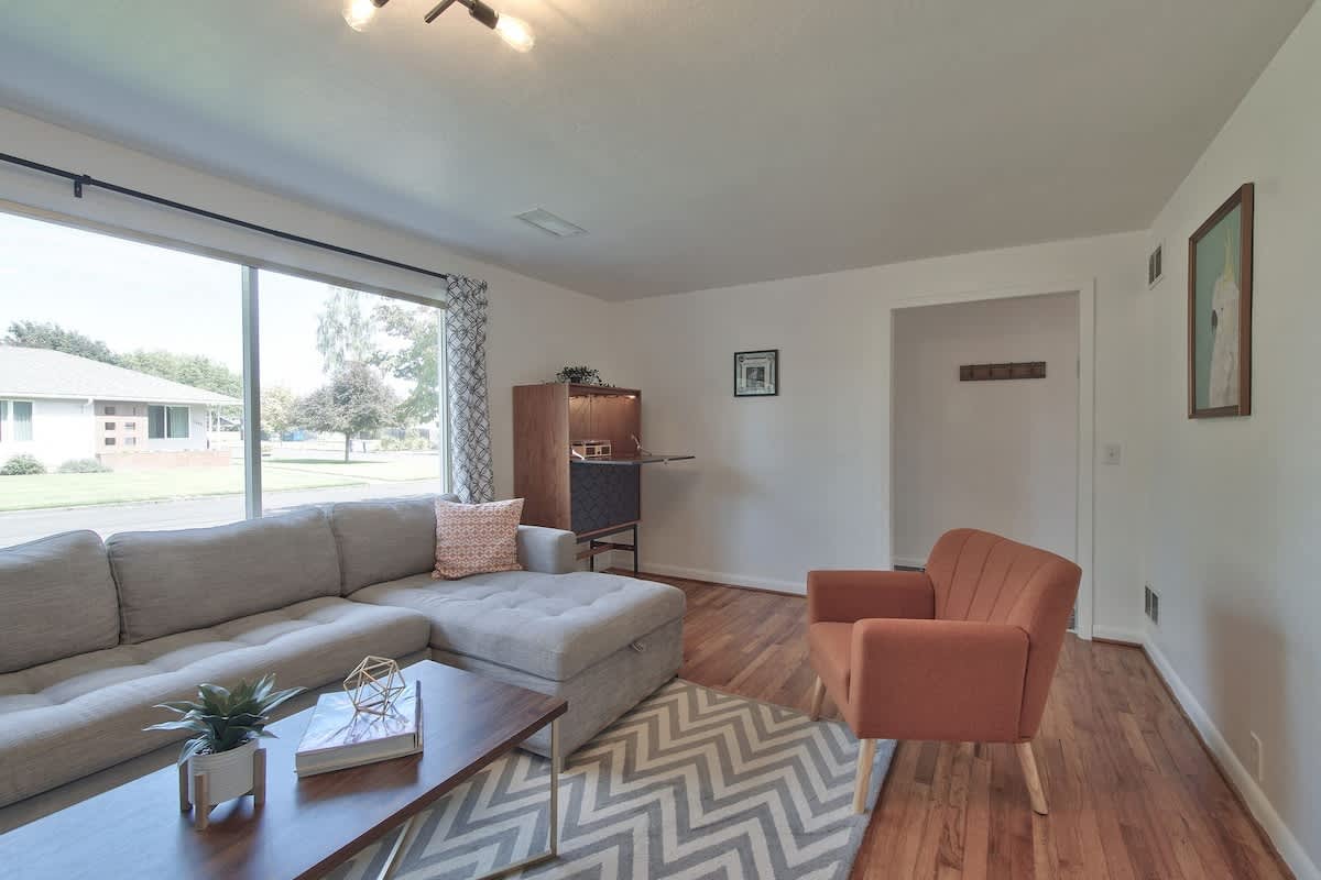 Modern living room featuring a large gray sectional sofa, a wooden coffee table, and a geometric rug. A wide window offers a view of the street and trees, with hardwood floors throughout.