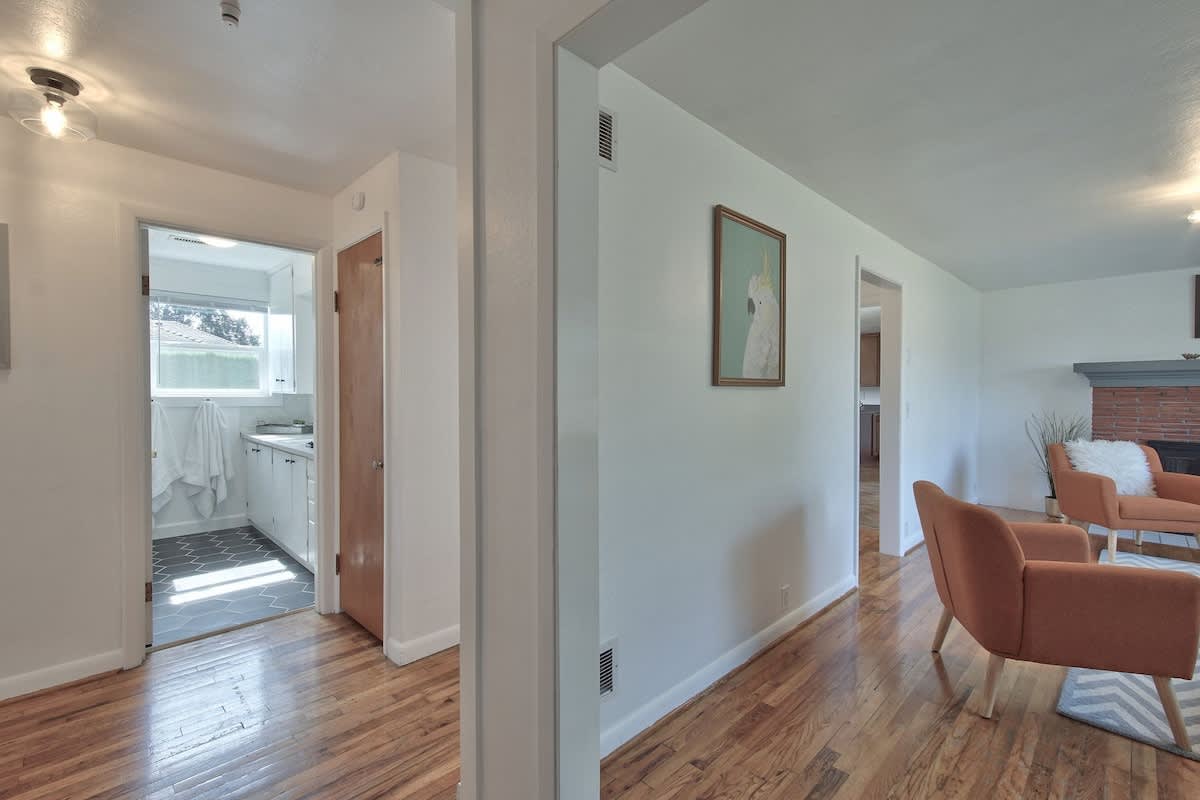 Bright hallway with polished hardwood floors, white walls, and doorways leading to the living room and another room, possibly a utility area or kitchen. A framed artwork hangs on the wall.