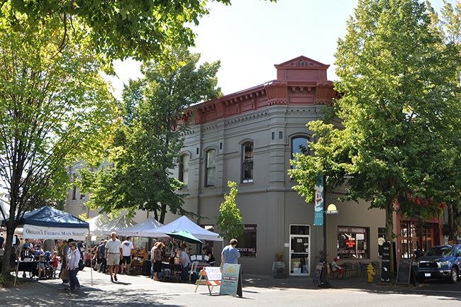 A vibrant outdoor market is set up on a sunny day, with numerous white tents and people browsing goods in front of a historic-style building with a red roof and grey facade. Mature green trees provide shade along the street.