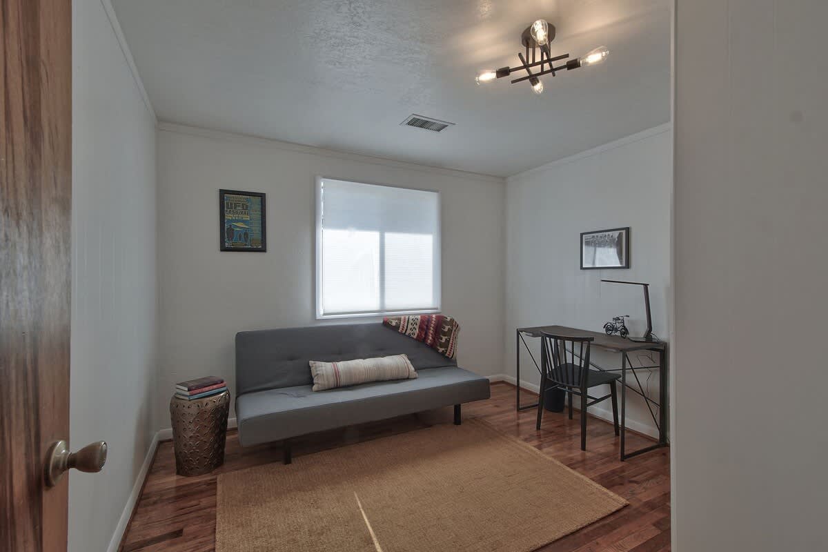 Dedicated office space featuring a gray futon, a modern desk with a chair, and a light brown area rug on hardwood floors. A window with blinds provides natural light, and framed artwork adorns the wall.