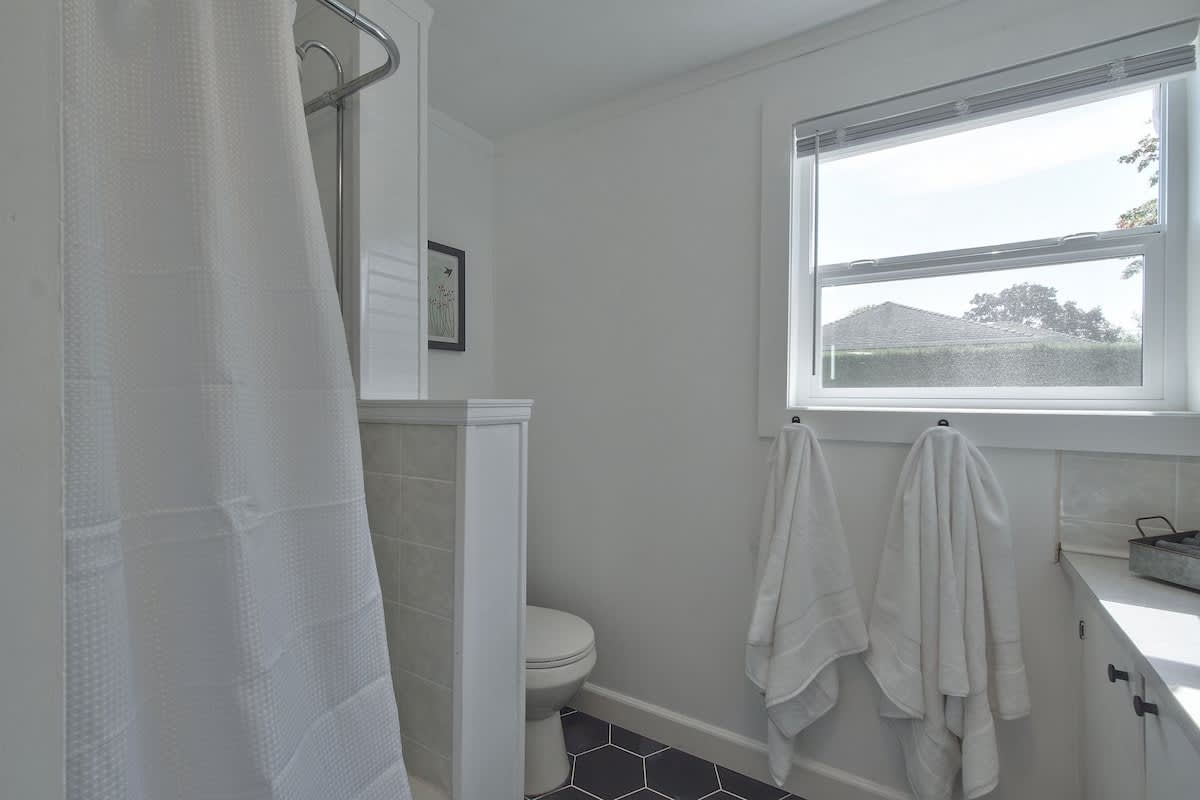 Clean white bathroom featuring a toilet, a shower with a white textured curtain, and a window with blinds. Dark hexagonal floor tiles contrast with the white walls and fixtures, and two white towels hang by the window.