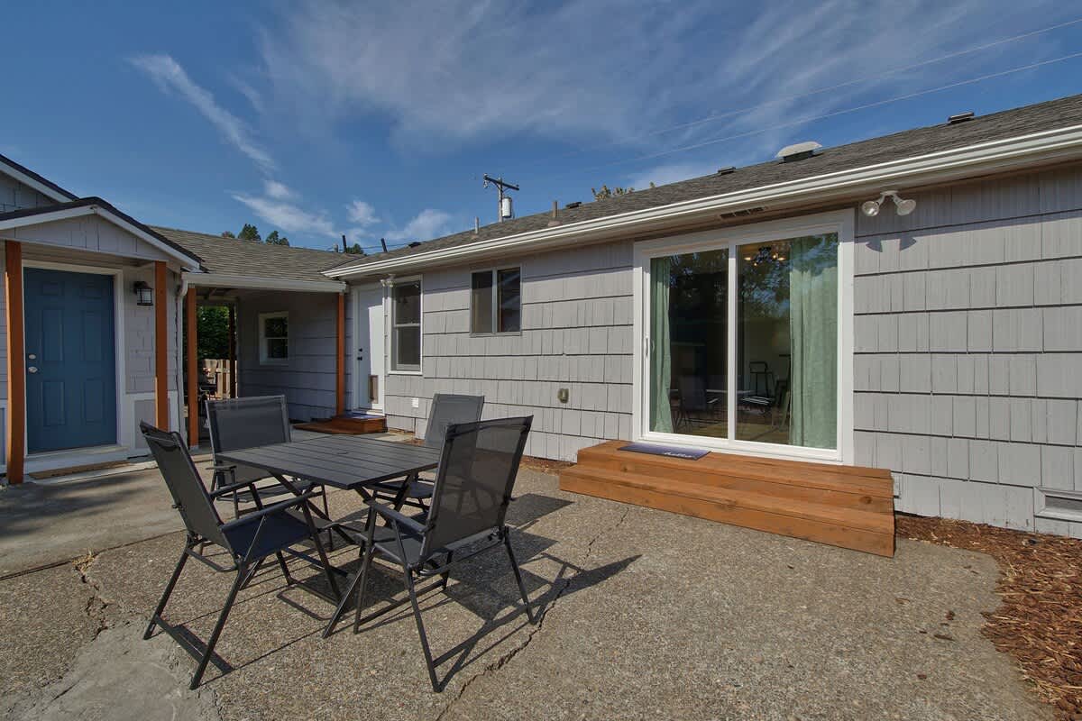 Outdoor concrete patio with a dark gray metal dining set for four, adjacent to the house with light gray siding and a sliding glass door leading indoors. A small wooden step leads to the door.