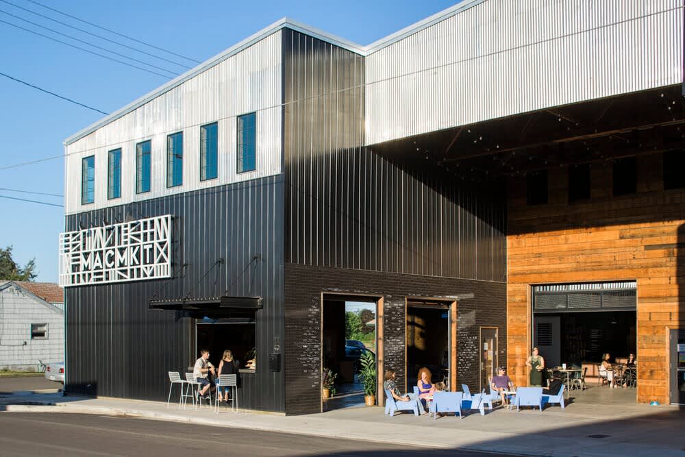 A modern commercial building features black and silver corrugated metal siding, large open doorways, and several groups of people enjoying outdoor seating on a sunny day. A distinctive "MACMTX" sign is visible on the black facade.