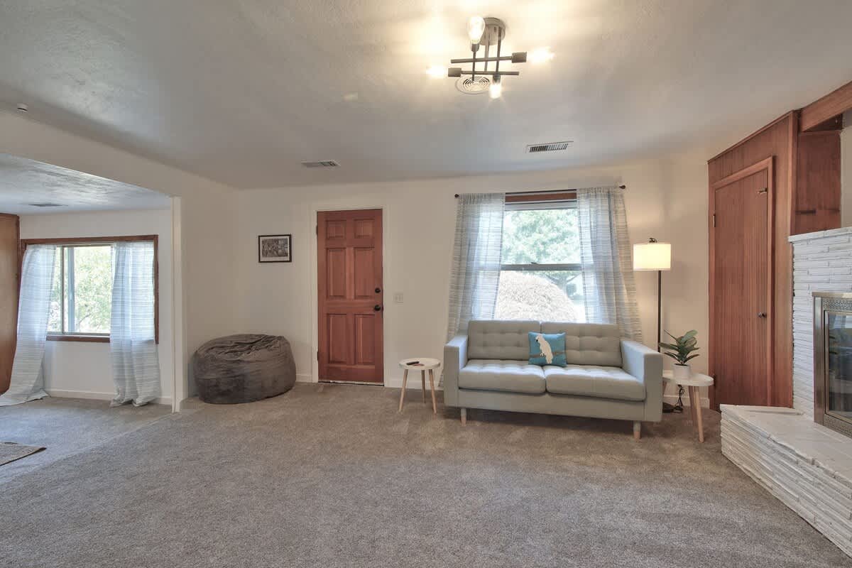 Bright living room with a light grey sofa and two small side tables, illuminated by a window with patterned curtains, and featuring a warm wooden front door and a large grey bean bag chair.