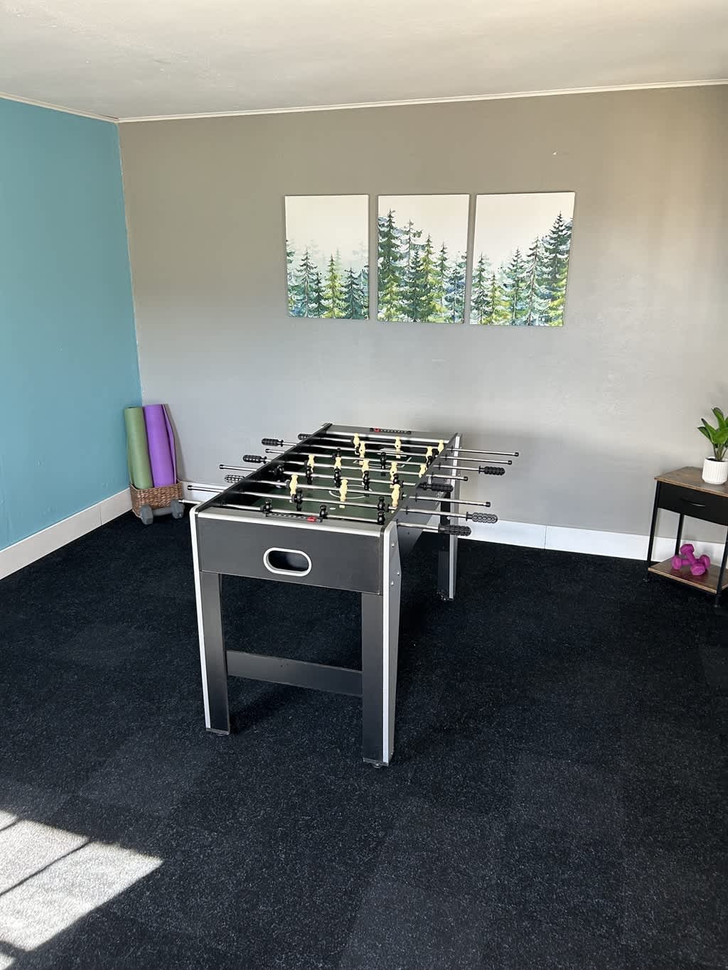 A black and white foosball table stands prominently on dark carpet in a game room, with three framed forest prints adorning the light gray wall.
