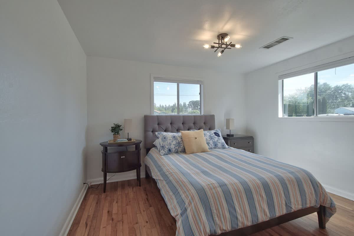 Full view of bedroom-2 with a queen-sized bed, gray tufted headboard, and striped bedding. Two dark wood nightstands with lamps flank the bed, and two windows provide ample natural light to the room with hardwood floors.