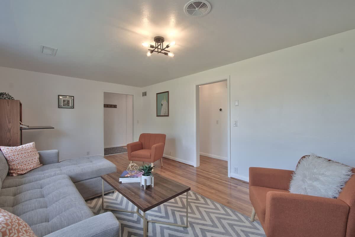 Inviting living room with a gray sectional sofa, two orange armchairs, and a geometric rug on hardwood floors. A hallway entrance is visible, and the room is well-lit by a ceiling fixture.