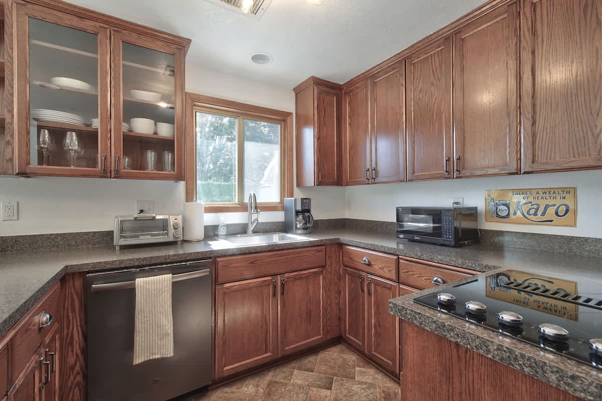A bright kitchen corner featuring a stainless steel sink beneath a window, surrounded by dark speckled countertops and ample dark wood cabinetry. Appliances include a toaster, coffee maker, and microwave, with dishes visible in glass-front cabinets.
