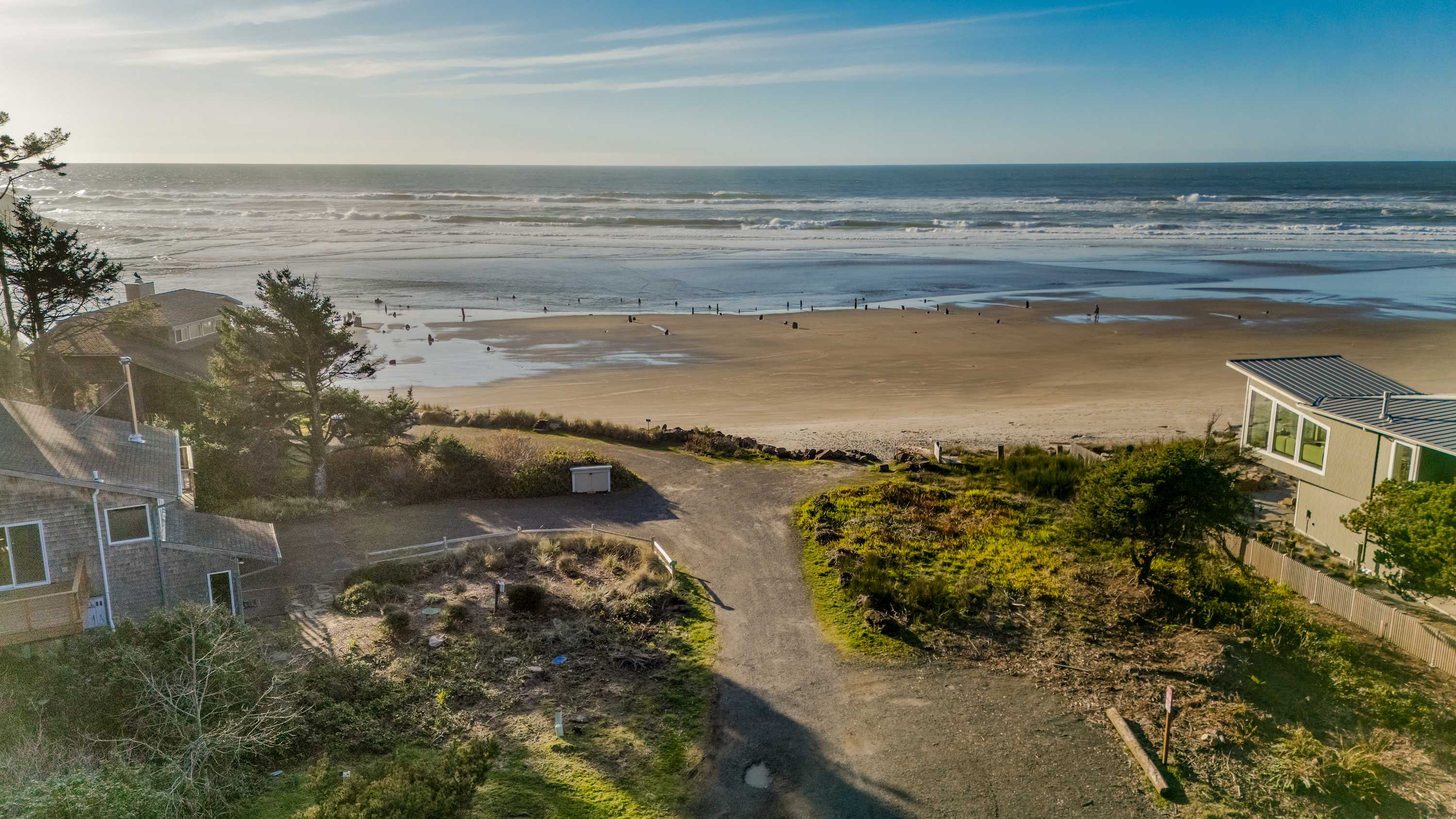 Aerial view of a gravel driveway leading to a house, with a wide sandy beach and the ocean in the background under a bright sky. The property is surrounded by green grass and sparse vegetation, with other houses nearby.