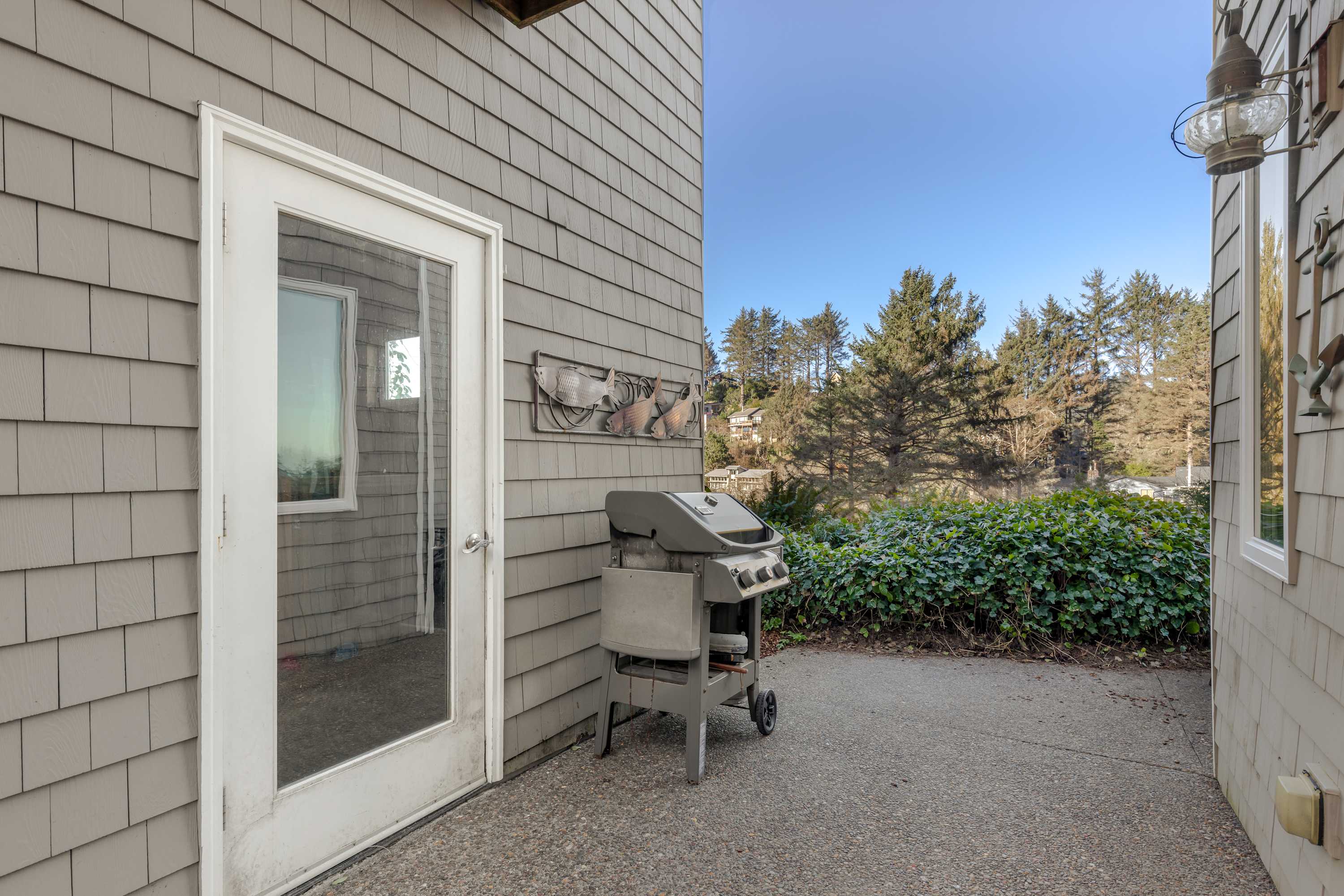 The exterior of the house showcases gray shingle siding and a white glass-paneled door leading to a concrete patio. A gas grill is positioned next to the door, with lush green foliage and trees in the background.
