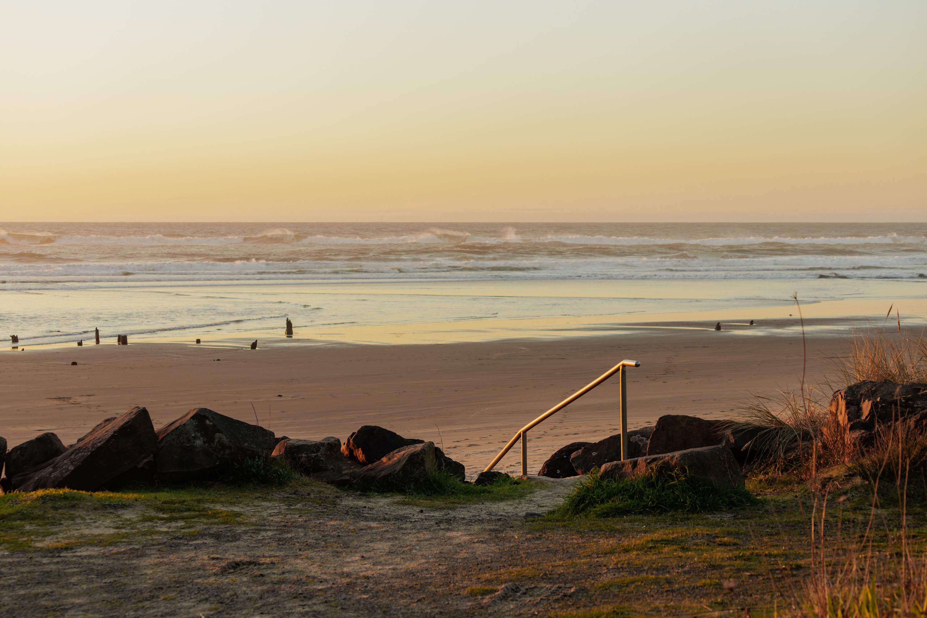 A view from a grassy dune overlooking a wide sandy beach and the ocean with breaking waves under a soft, golden sunset sky, featuring a metal handrail leading down to the sand.