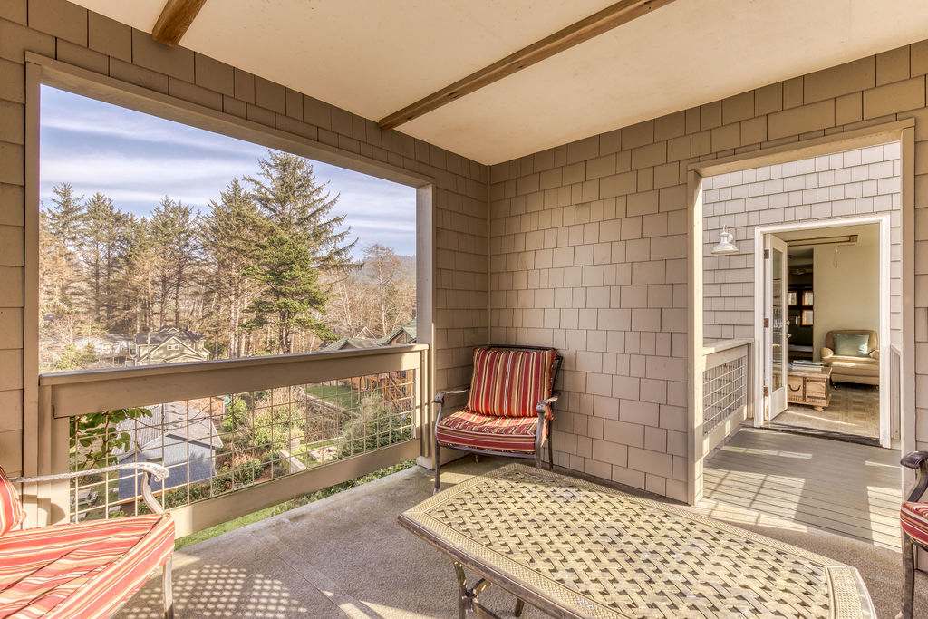 A covered balcony area within the lookout tower, furnished with two striped outdoor armchairs and a small table, offering views of surrounding trees and distant houses. The space features light brown shingle walls and a wooden railing.
