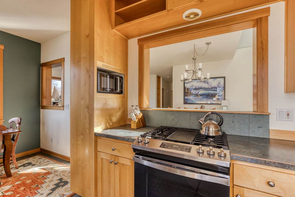 Kitchen view showing a gas range with a griddle, a microwave built into light wood cabinetry, and a pass-through window connecting to the dining area, with dark countertops and a tiled backsplash.