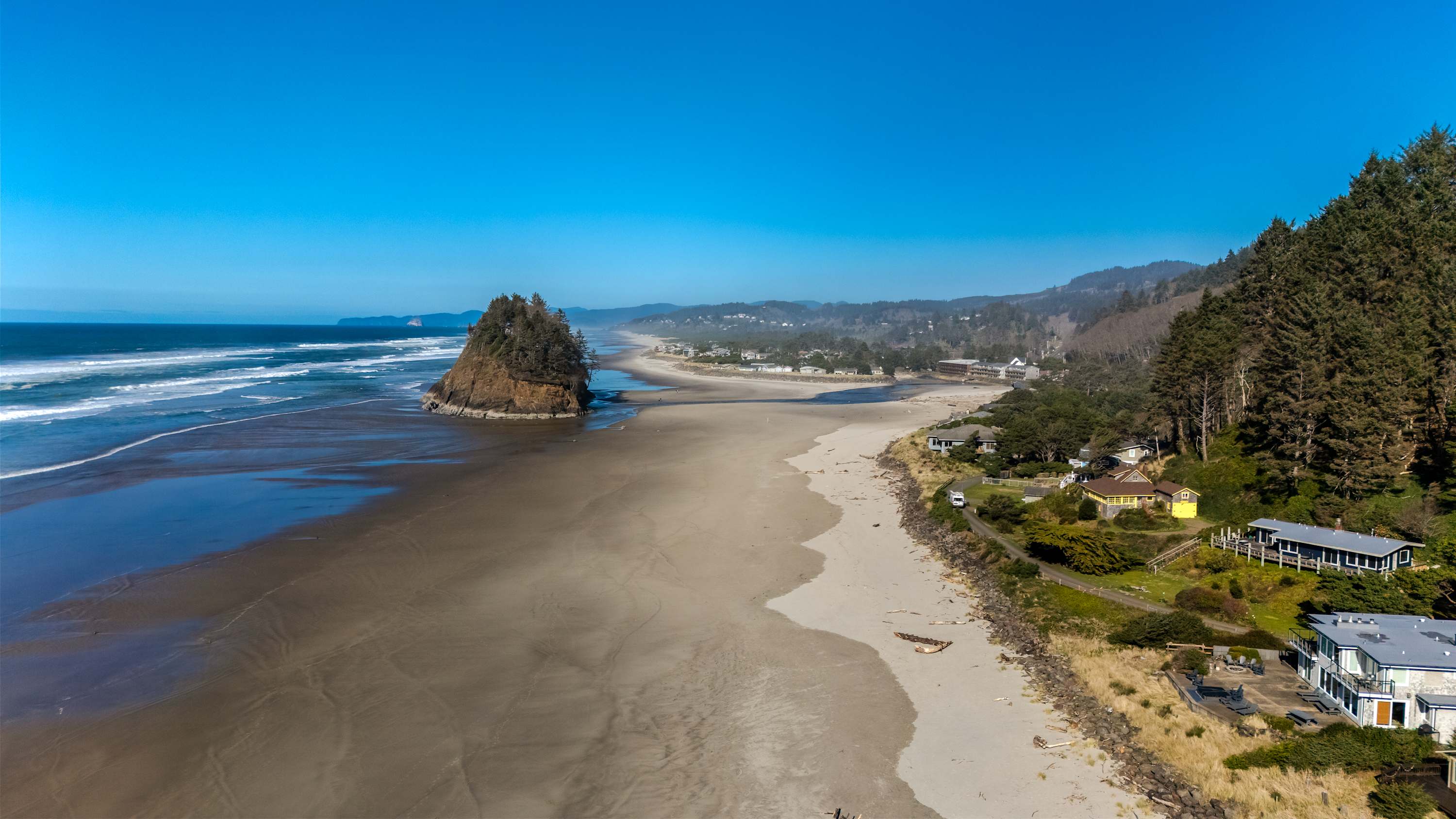 Panoramic aerial view of a long sandy beach stretching along the coastline, with a prominent sea stack (Proposal Rock) in the distance. Houses are visible nestled among evergreen trees on the adjacent hillside, under a bright blue sky.